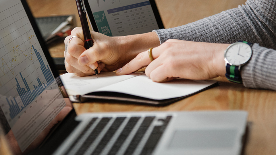 Two individuals working on laptops and one writing in a notebook.